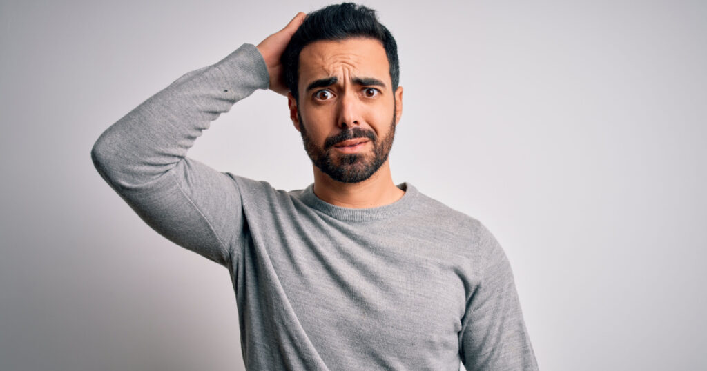Young handsome man with beard wearing casual sweater standing over white background confuse and wonder about question. Uncertain with doubt, thinking with hand on head. Pensive concept.

