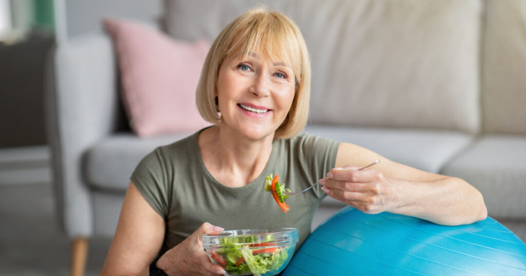 Exercise and healthy diet concept. Senior woman with fitball eating fresh vegetable salad at home. Mature Caucasian lady having veggies after sports training, keeping weight loss diet
