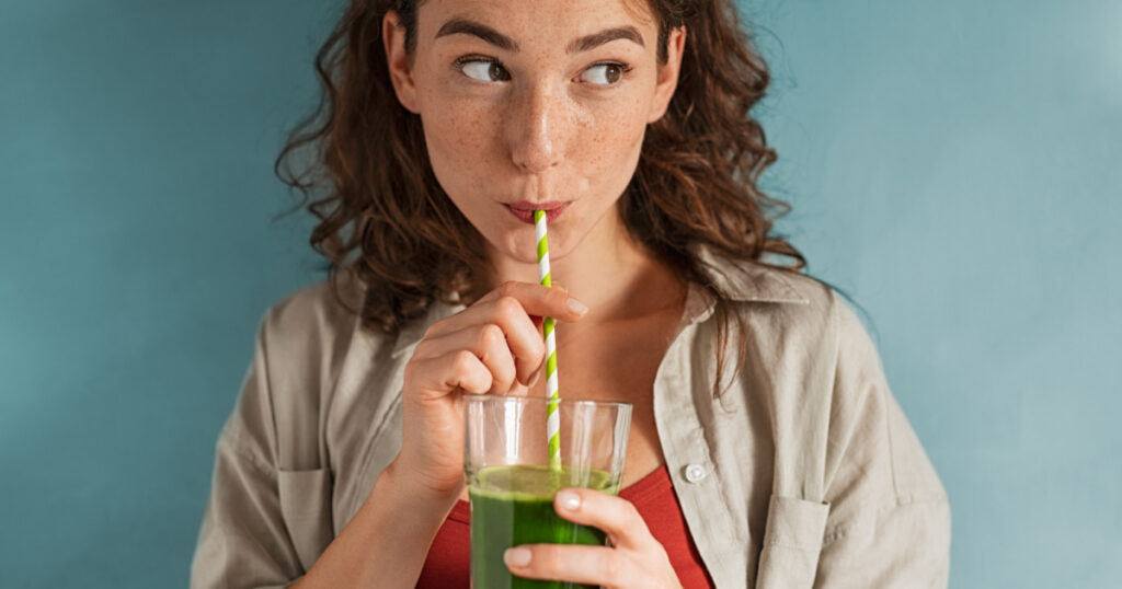 Beautiful woman drinking an organic green smoothie. Fit young woman drinking detox juice using paper straw isolated against blue background. Healthy girl enjoy detox drink and looking away.
