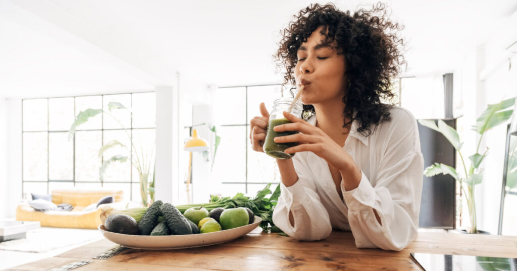 Young african american woman drinking green juice with reusable bamboo straw in loft apartment. Home concept. Healthy lifestyle concept. Copy space
