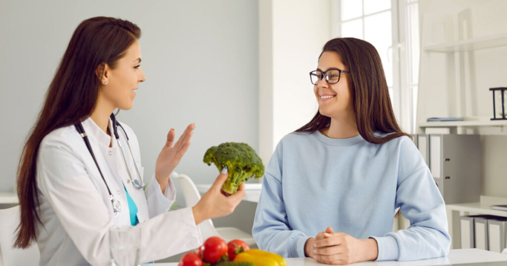 Female nutritionist tells young woman about benefits of eating vegetables during consultation. Smiling woman listens to qualified healthy food expert who holds broccoli in her hands and speaks.