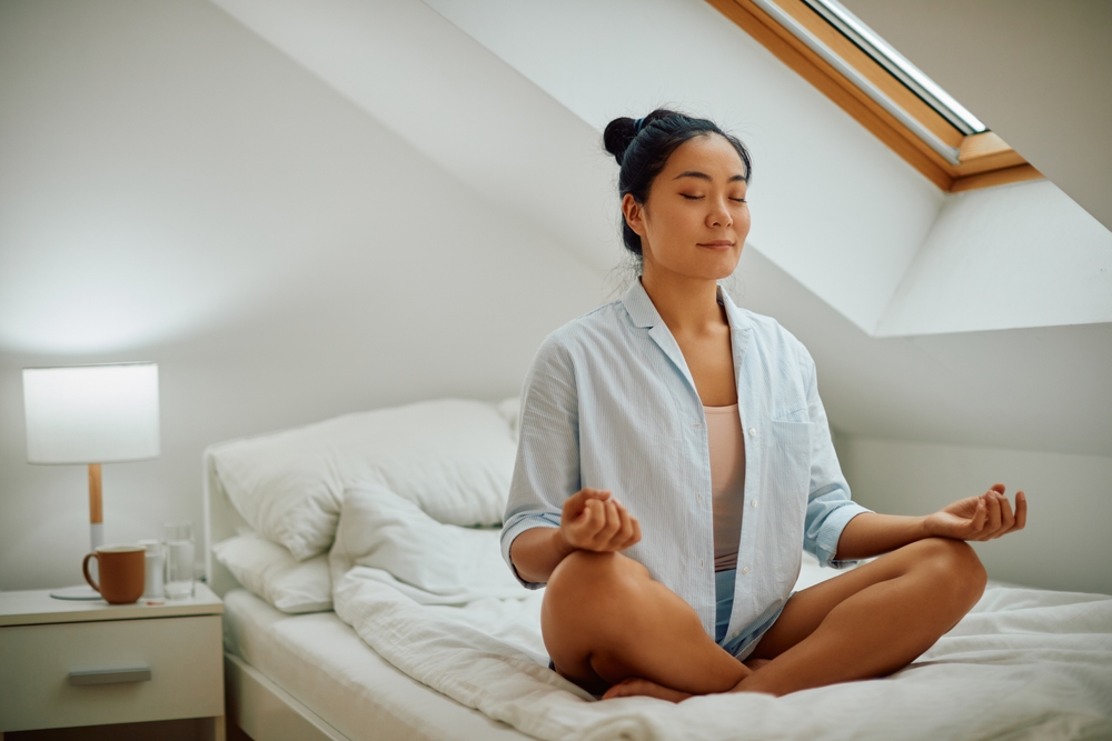 Young Asian woman in lotus pose doing breathing exercise while practicing Yoga in her bedroom. Copy space.

