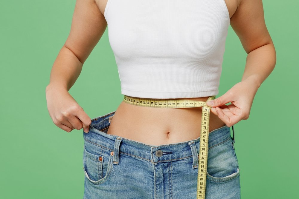 Cropped young woman in white clothes show loose pants after weightloss hold measure tape on waist isolated on plain pastel green background. Proper nutrition healthy fast food unhealthy choice concept
