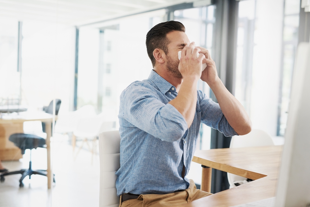 Business man, blowing nose and allergy in office with flu, virus and tissues for healthcare at desk. Young businessman, allergies or sick with hayfever, toilet paper and cleaning mucus in workplace