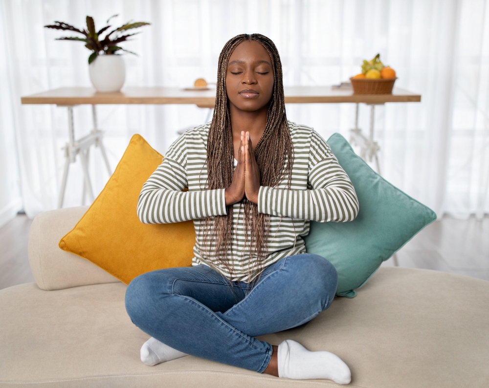 Peaceful pretty young black woman with long braids in casual outfit sitting on couch in lotus positon, meditating with closed eyes, home interior, copy space. Mindfullness, stress relief concept

