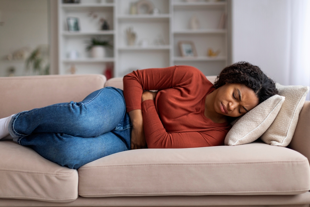 Young black woman clutching her abdomen in discomfort at home, african american female suffering from menstrual pain or stomach issues, lying on sofa in her living room, free space
