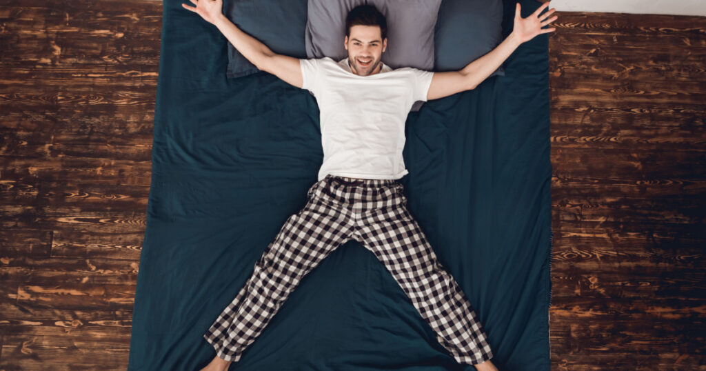 Top view. Adult cheerful man shows star, placing arms and legs aside on bed. Rest at home. Home leisure. Sleep poses. Smiling young man shows starfish.