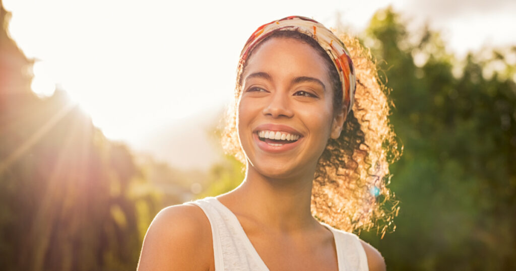 Portrait of beautiful african american woman smiling and looking away at park during sunset. Outdoor portrait of a smiling black girl. Happy cheerful girl laughing at park with colored hair band.