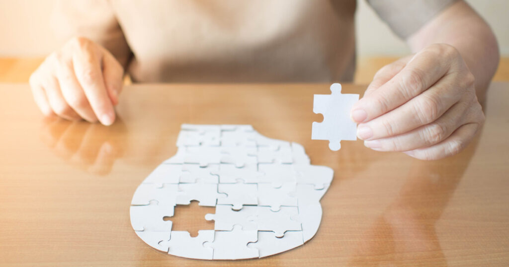 Elderly woman hands holding missing white jigsaw puzzle piece down into the place as a human head brain shape. Creative idea for memory loss, dementia, Alzheimer's disease and mental health concept.