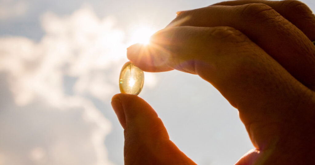 Vitamin D keeps you healthy while lack of sun. Yellow soft shell D-vitamin capsule against sun and blue sky on sunny day. Cure concept.