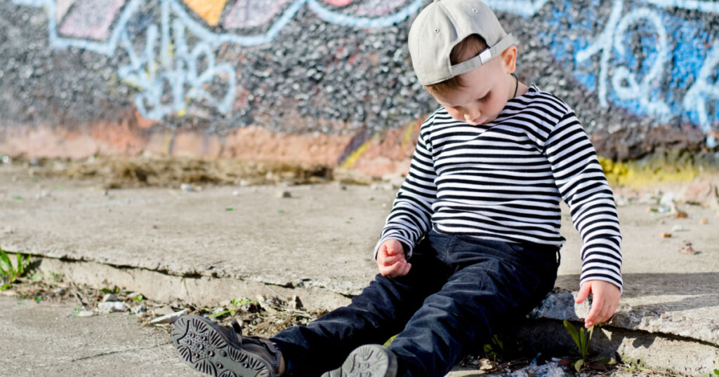 Lonely little boy sitting on the sidewalk playing with stones in the gutter in front of a brightly coloured wall covered in graffiti