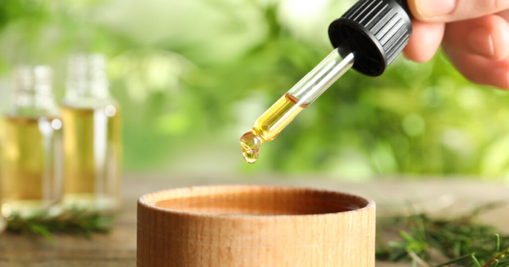Woman dripping natural tea tree oil in bowl on table, closeup