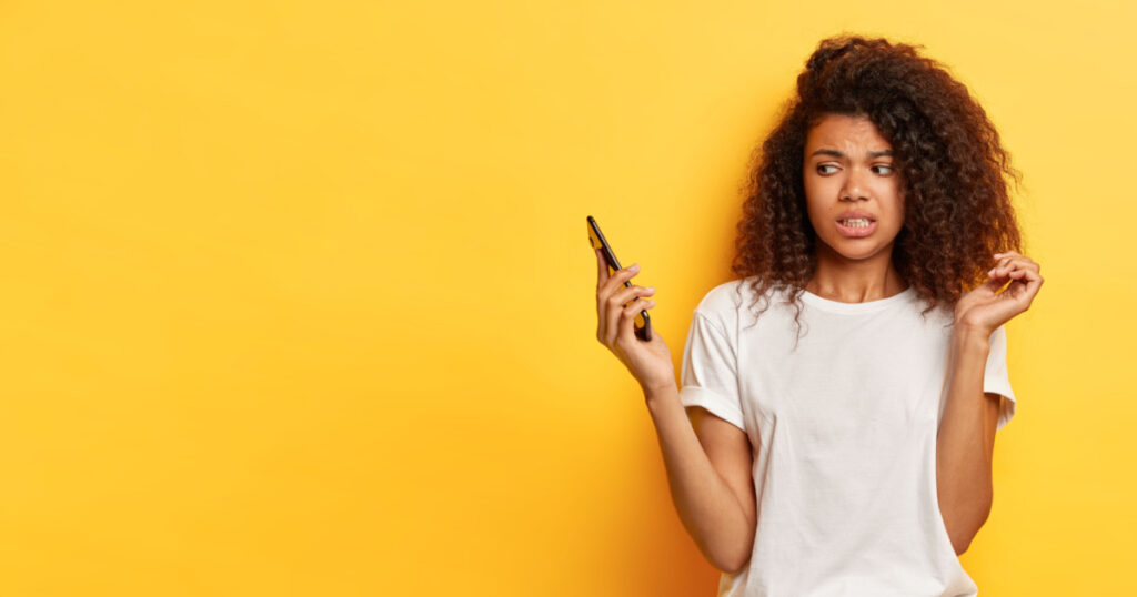 Beautiful female with frizzy Afro hairstyle, looks in displeasure at cell phone, receives call from former boyfriend, holds mobile phone on distance, ignores communication, afraids of something