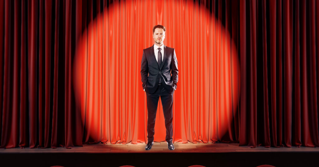 Attractive businessman standing on red stage in spotlighting. Star, show, speech and presentation concept.
