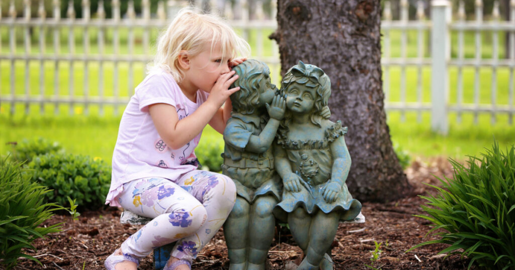 A cute little girl is sitting on a bench, whispering a secret as she plays with two little garden statue friends.