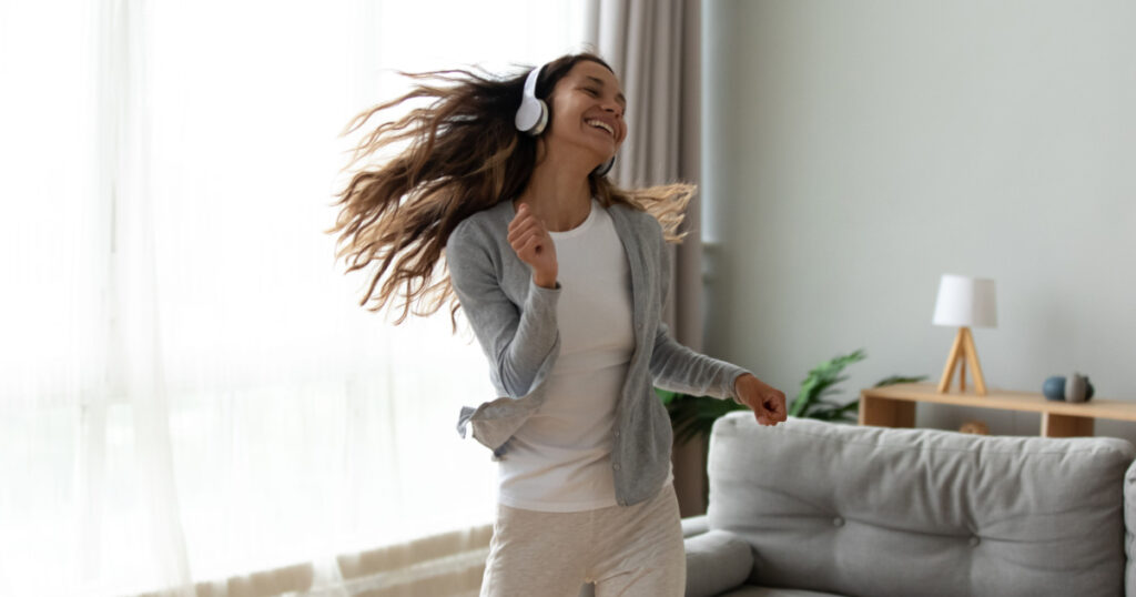 Overjoyed millennial girl wearing headphones have fun moving listening to music relax in living room, happy young woman in earphones dance enjoy leisure weekend at home, stress free concept