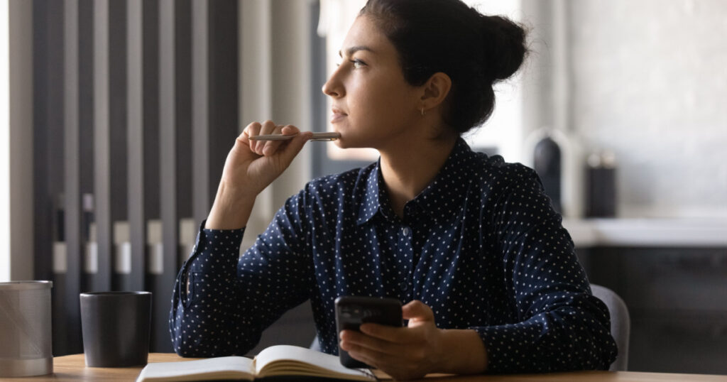 Pensive young hindu female study by desk using mobile internet distracted from making notes create new idea. Thoughtful mixed race woman looking aside of phone screen pondering planning future work