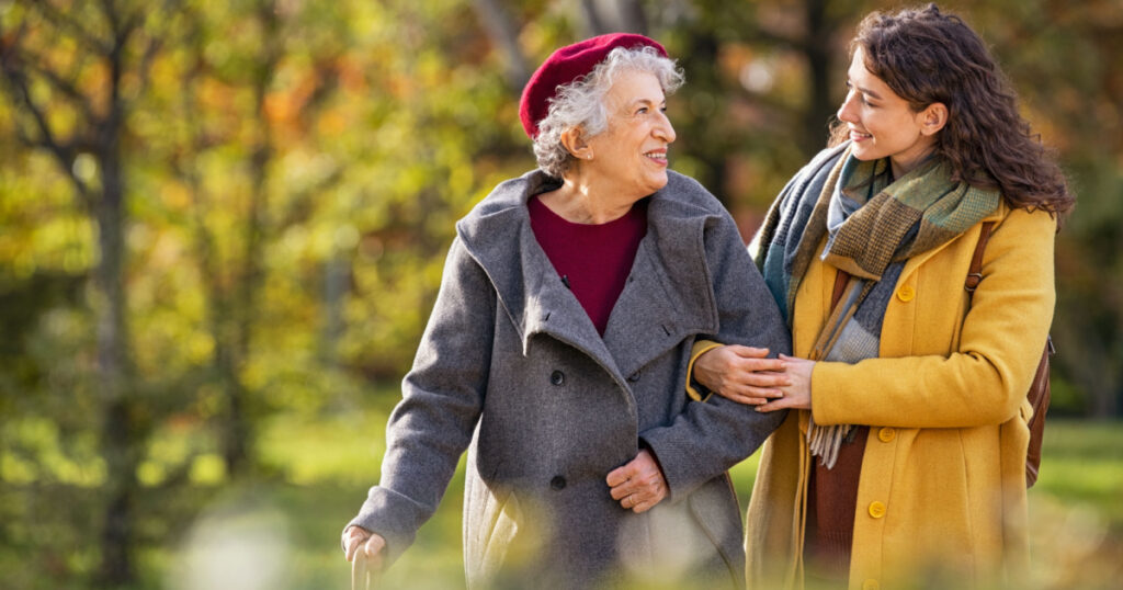 Young woman in park wearing winter clothing walking with old grandmother. Smiling lovely caregiver and senior lady walking in park during autumn and looking at each other with copy space.
