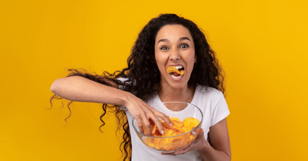 Binge Eating Concept. Portrait Of Funny Hungry Woman Enjoying Delicious Potato Crisps Holding Glass Bowl, Posing With Chips In Mouth Isolated Over Yellow Orange Studio Wall. Junk Meal Addiction
