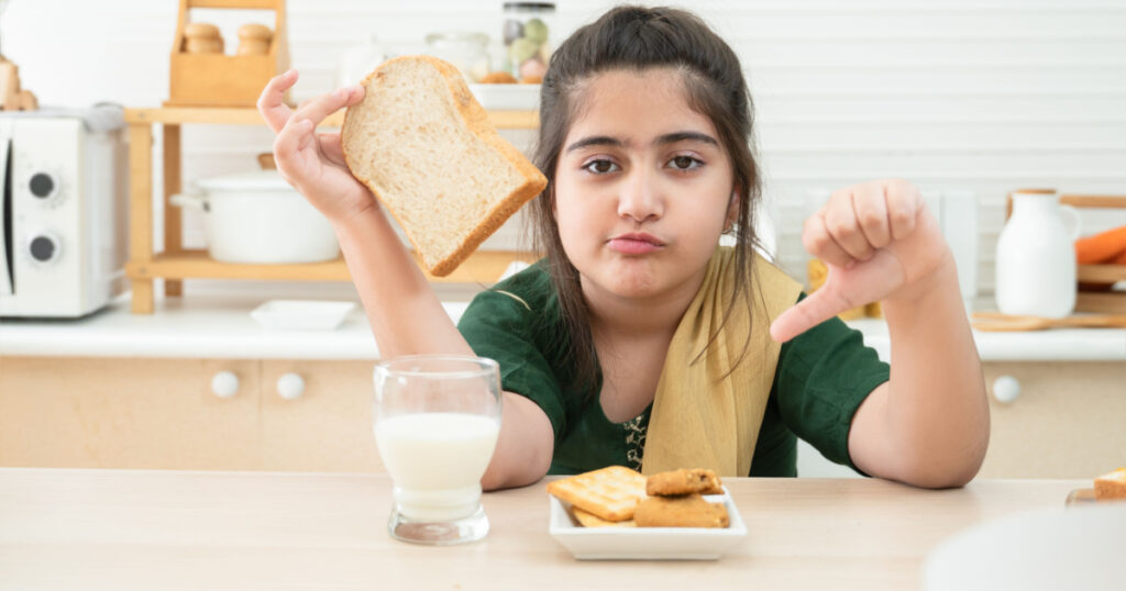 Little cute Indian kid girl dislike breakfast holding sliced bread and thumbs down while having breakfast with a glass of milk, crackers and cookies in kitchen at home
