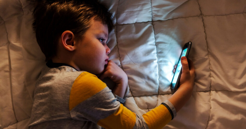Cute little boy using a smartphone. Kid playing with mobile phone, lying on a bed. Freetime. Technology and internet concept. Smiling toddler laying on bed and looking at smartphone. Light reflectio