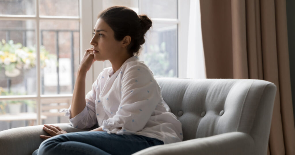 Sad upset young Indian woman thinking while sit on armchair and staring out window. Personal troubles and break up, goes through difficult life situation, search solution, ponders seated alone at home