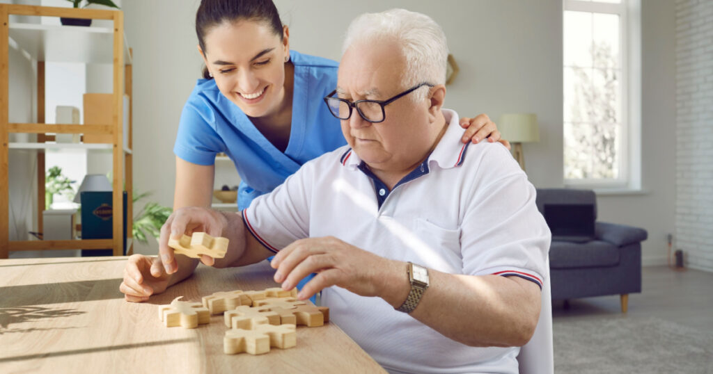 Nurse in retirement home helping old male patient with board game. Senior man with cognitive disorder sitting at table in geriatric clinic, playing games and thinking. Dementia, Alzheimers, care