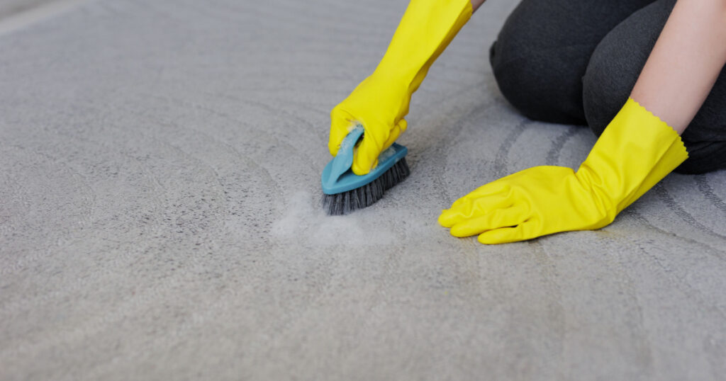 close up of female hands in yellow rubber gloves cleaning carpet with brush and foam, copy space over grey carpet
