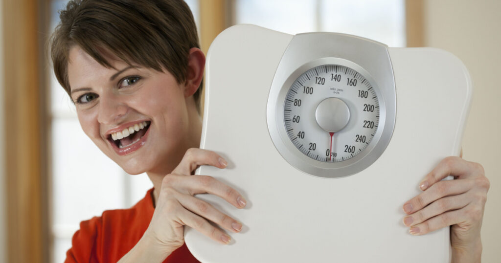 Attractive young woman holds a bathroom scale up while smiling at the camera. Horizontal shot.