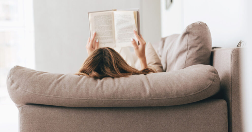 Back view of woman lying on sofa and reading book at home