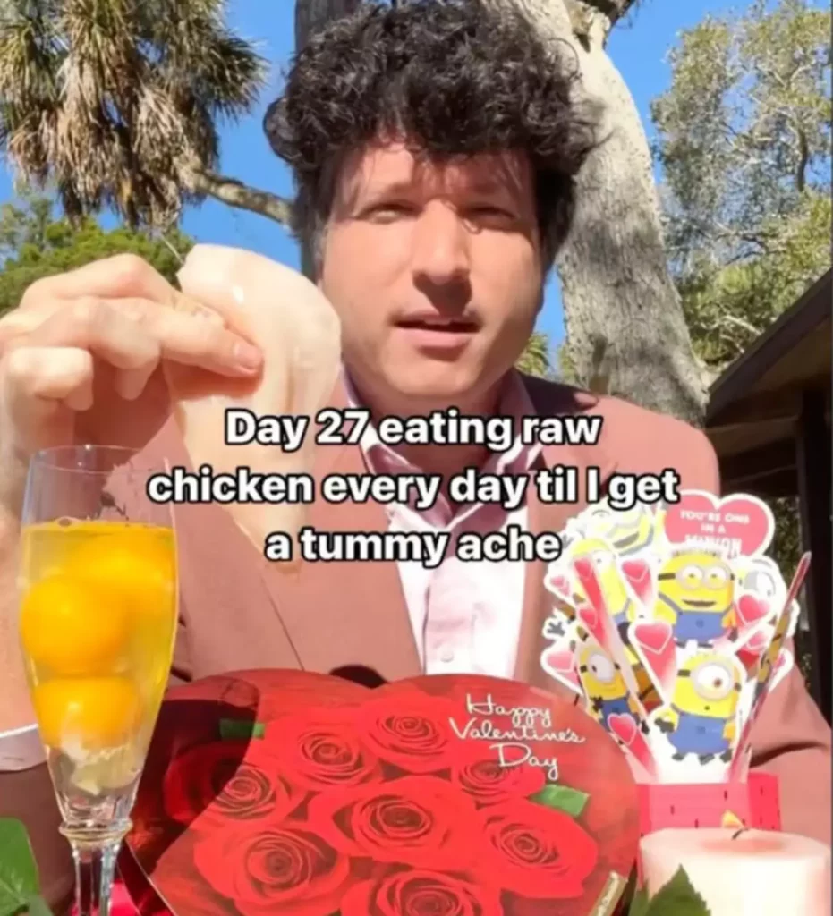 Man with raw chicken and eggs, trees in the background and valentines treats scattered on the table.
