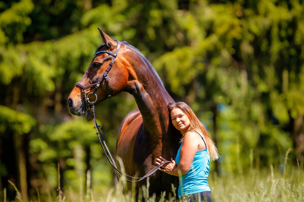 Horse stands with a young woman in a meadow, horse looks attentively to the right and the woman snuggles up to the horse's neck.