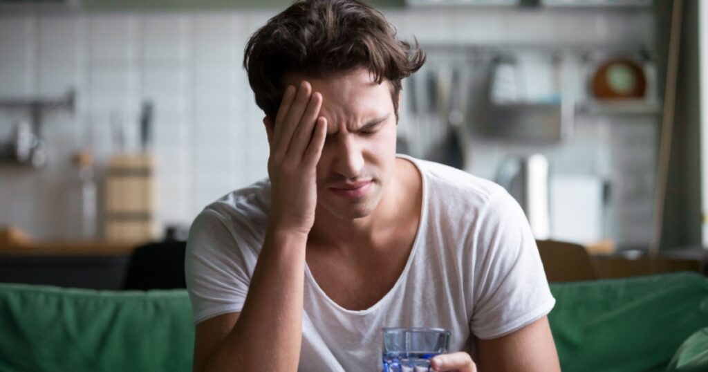 Young man suffering from strong headache or migraine sitting with glass of water in the kitchen, millennial guy feeling intoxication and pain touching aching head, morning after hangover concept