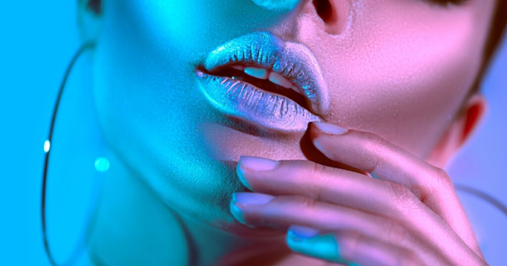 High Fashion model metallic silver lips woman in colorful bright neon blue and purple lights posing in studio, beautiful girl, trendy glowing make-up, colorful metal make up. Glitter Vivid neon makeup