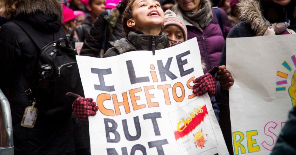 Young girl shouts into the crowd while holding a sign "I Like Cheetos but Not This One" while marching in the NYC Women's March - New York, NY, USA January 1/19/2019 Women's March
