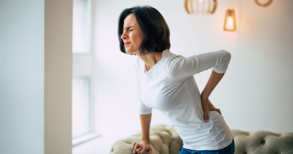 Chronic back pain. Adult woman is holding her lower back, while standing and suffering from unbearable pain.