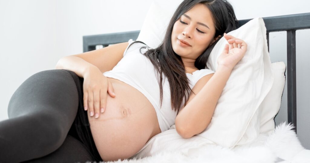 Beautiful Asian pregnant woman lie with relax position and use one hand touch her belly on bed. Concept of good healthy activity for mother and support growth of baby in womb of people.
