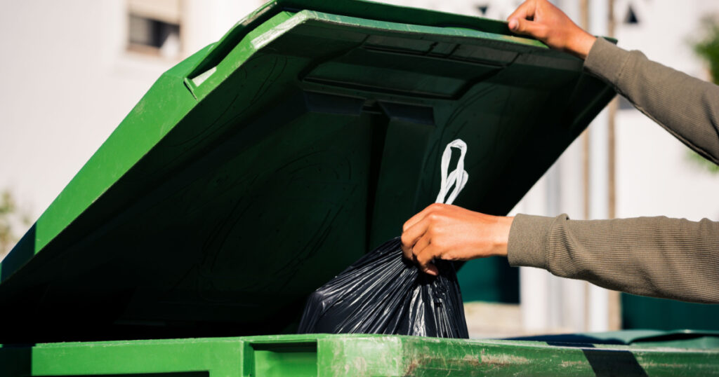 Man throwing out black eco-friendly recyclable trash bag in to big plastic green garbage container. Take out the trash