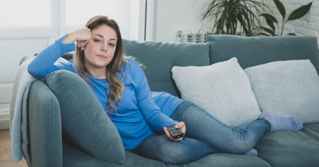 Bored woman watching TV helpless in self Isolation at home during mandatory lockdown due to coronavirus outbreak. Young upset woman on sofa using control remote zapping bored of TV and sedentary life.
