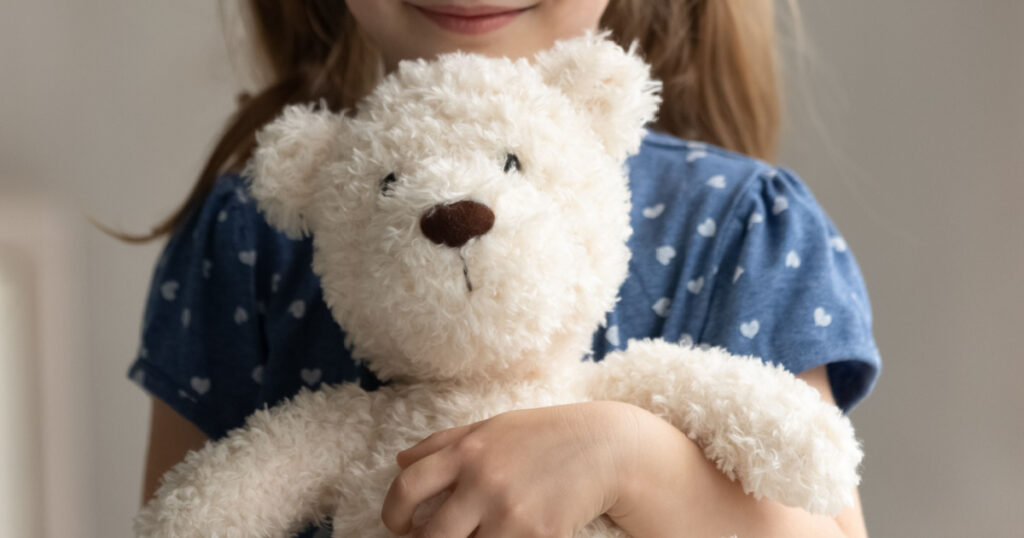 Close up of teddy bear in arms of child. Happy smiling little girl kid holding, hugging toy, playing with plush soft friend. Cropped portrait. Childhood, preschooler, playtime, childcare concept