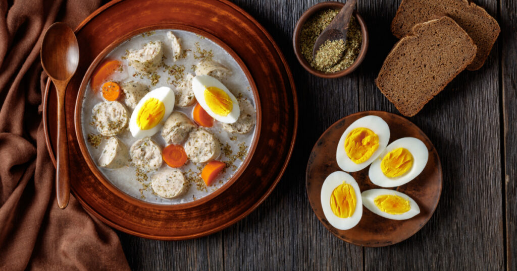 Zurek polish fermented rye soup with traditional polish white kielbasa or sausage with marjoram, hard-boiled eggs served on a clay bowl with a wooden spoon, on a dark wooden background, top view
