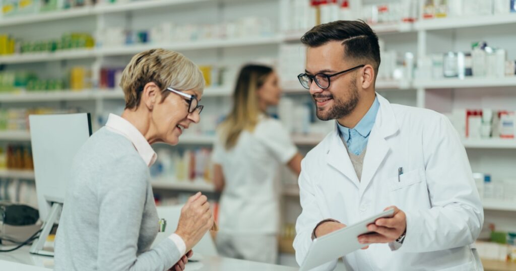 Young male pharmacist giving prescription medications to senior female customer in a pharmacy with female pharmacist in the background