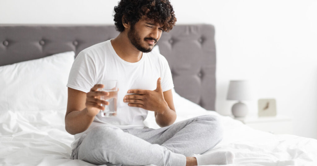 Unhappy millennial indian guy suffering from heartburn, waking up in morning, sitting on bed and holding glass of water, wearing pajamas, copy space. Acid reflux, pain in stomach concept
