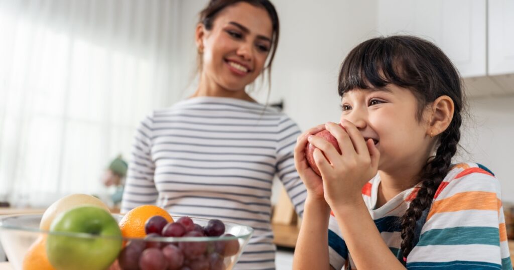 Asian mother teach and motivate young girl child eat healthy fruit. Adorable little kid child feel happy and relax, enjoy eat apple healthy foods for health care and wellness in kitchen at home.