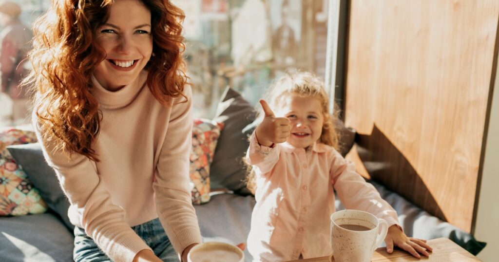 Smiling happy European woman with red wavy hair sitting in cafe in sunny day with her little daughter. Portrait of gorgeous stylish young mom with brown hair and little girl looking away.