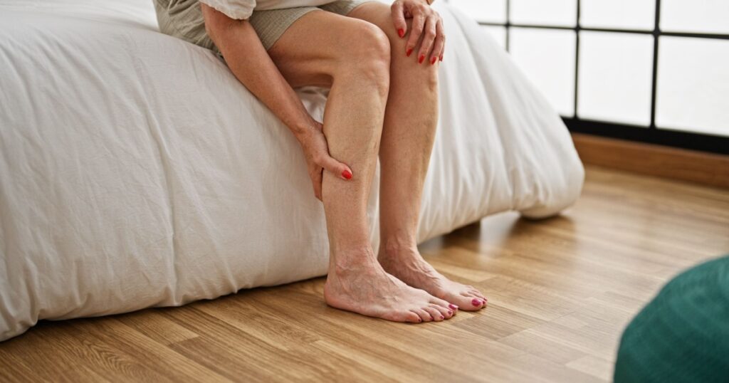 Middle age woman sitting on bed tired massaging legs at bedroom