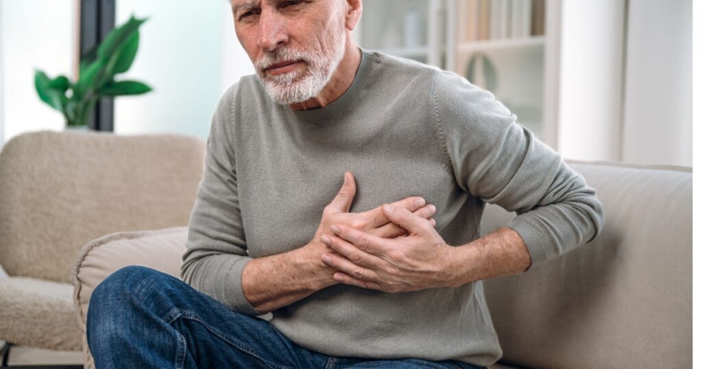 mature man feeling unwell, cardiac pain in chest, sitting on couch at home and need medical help. senior male has heart attack symptom. healthcare and medicare concept