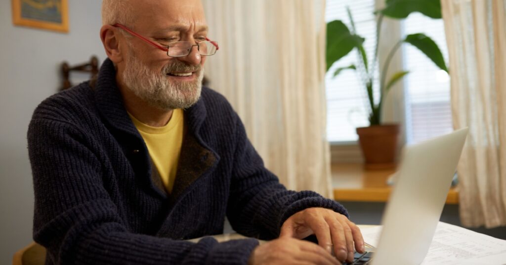 Side view of senior male professor in glasses and cardigan working on laptop sitting at table next to window at home, teaching online. Elderly man with beard chatting wit his friends on computer
