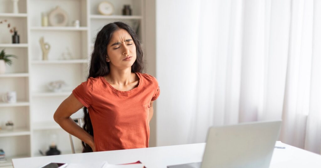 Weary young indian woman working on laptop at home office, sitting at desk with closed eyes, rubbing her lower back, suffering from office syndrome, copy space. Burnout at wok, overworking