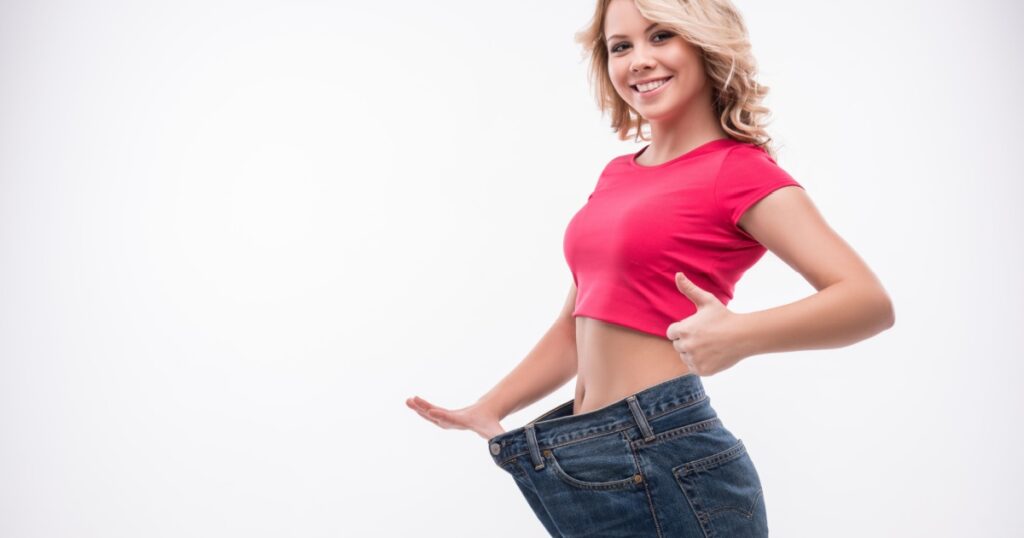 Full-length portrait of attractive slim young smiling woman in big jeans showing successful weight loss with her thumb up, isolated on white background